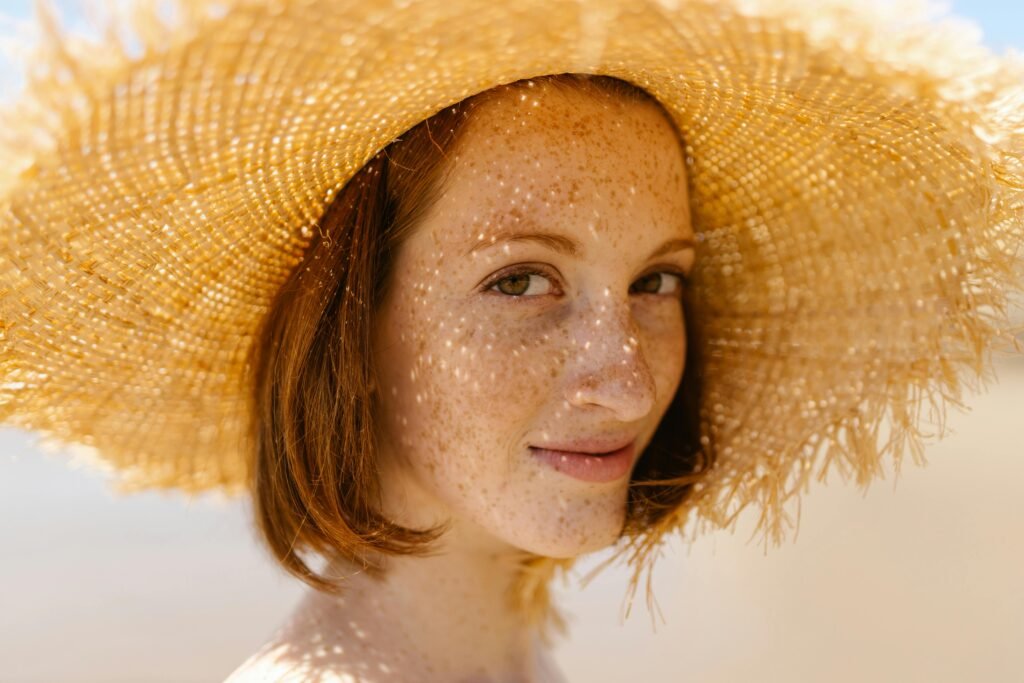 Close-up of a smiling woman with freckles wearing a straw hat outdoors.