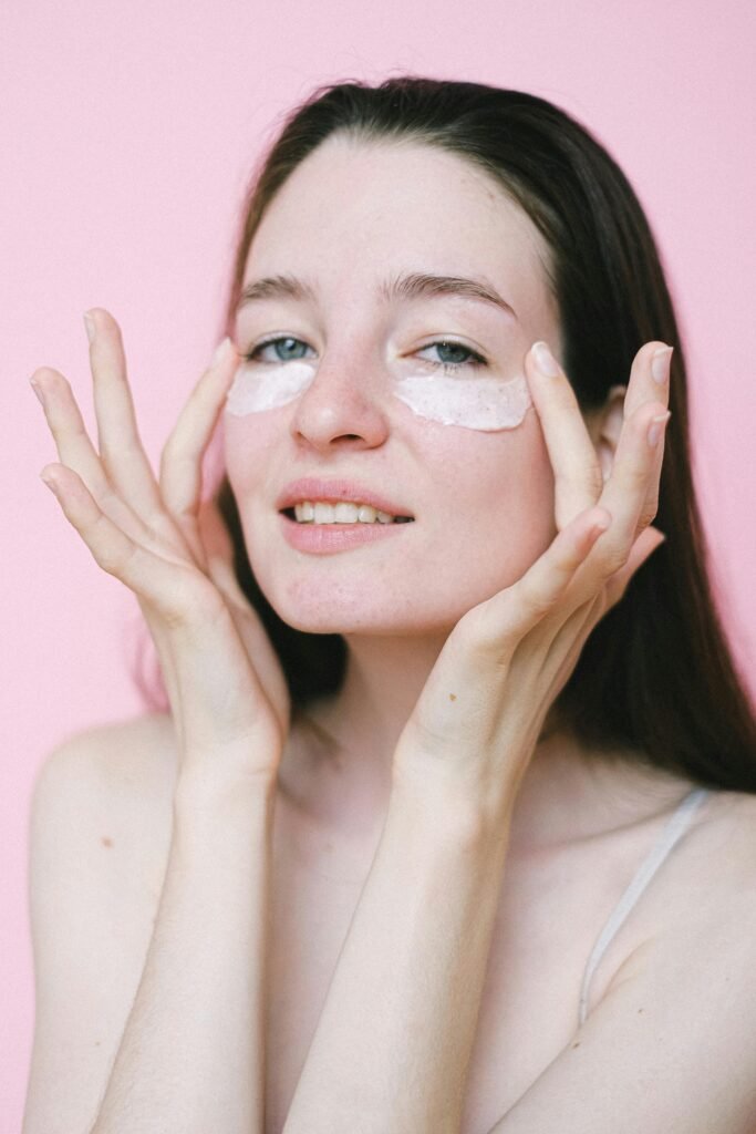 Cheerful female with naked shoulders smearing moisturizing cream on face and looking at camera against pink background