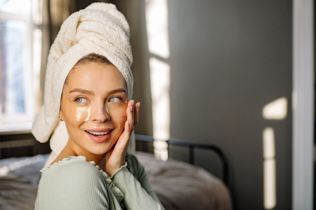 A woman with a towel wrapped around her head applying under-eye masks in a sunlit room.